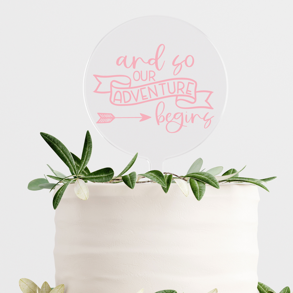 Circular clear cake topper with "and so our adventure begins" written in pink on a simple white cake