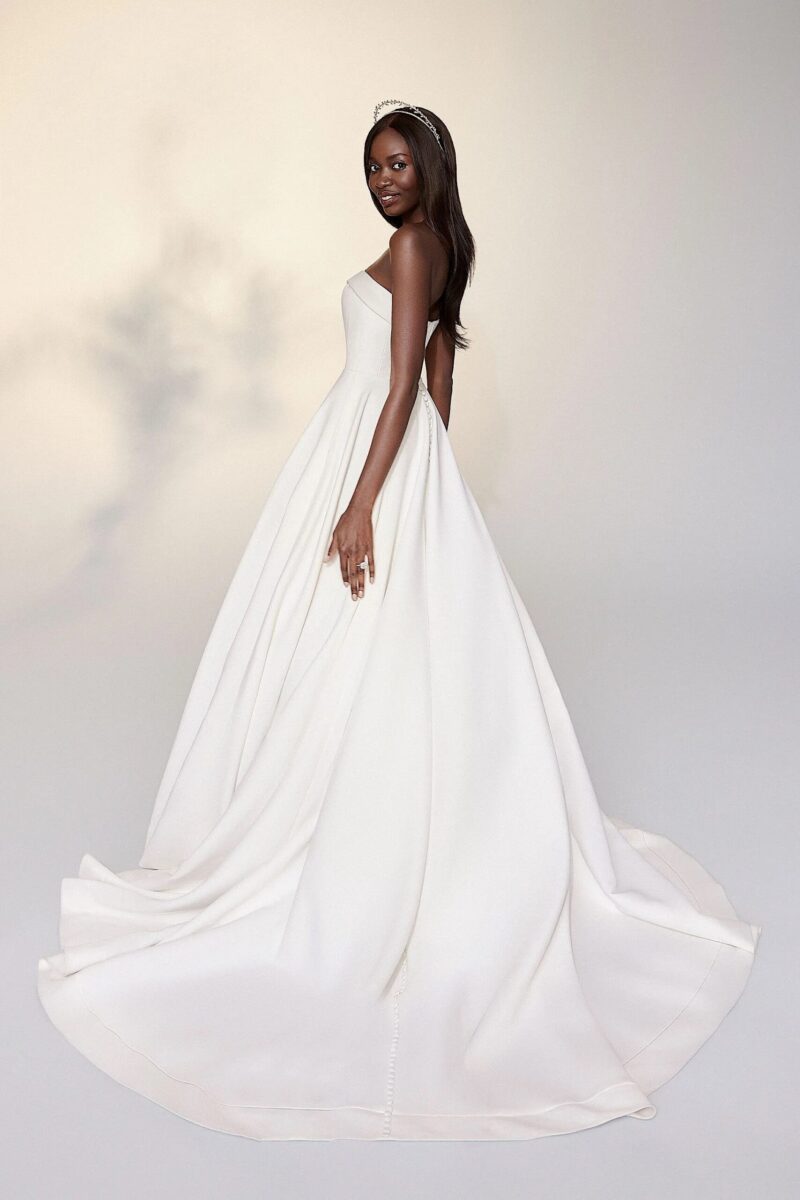 A woman modeling a strapless white wedding gown with a full, flowing A-line skirt and train against a soft light gray studio background.