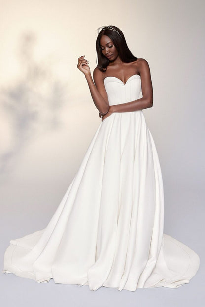 A woman modeling a strapless white wedding gown with a sweetheart neckline, full A-line skirt, and a short train against a soft light gray studio background.