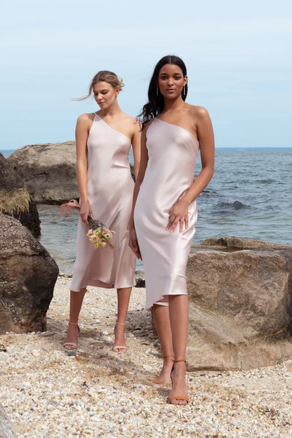 Two women in one-shoulder pink dresses standing on a rocky beach.
