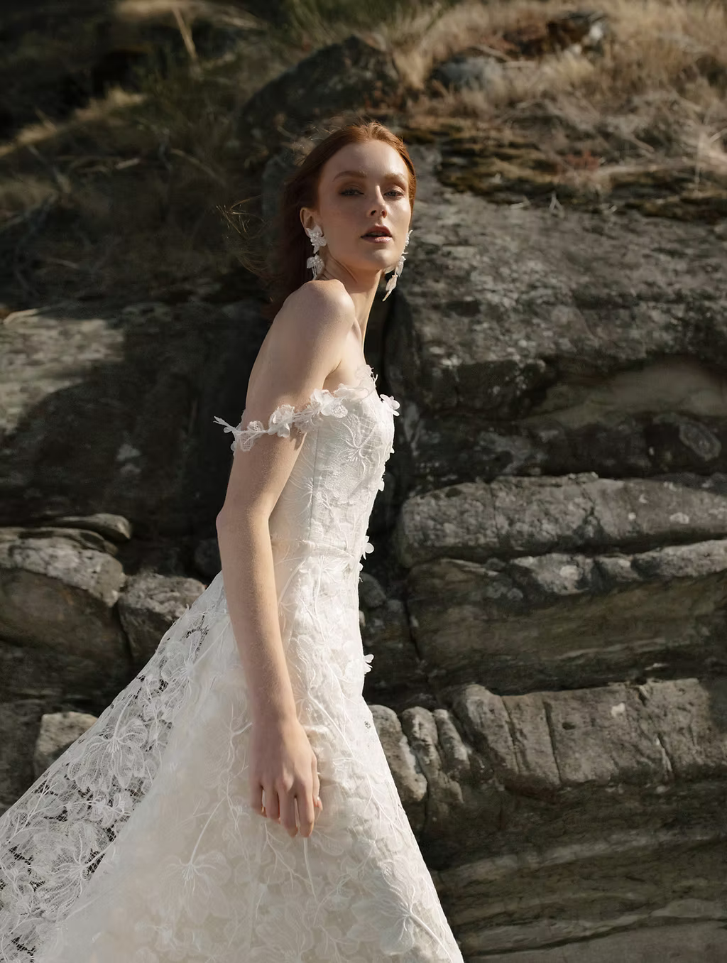 A woman wearing a strapless white lace wedding gown, suqare neck agnes by ouma, stands outdoors against a rocky cliffside background.
