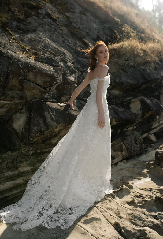 A woman wearing a strapless white lace wedding gown, Agnes by Ouma Caanadian Bridal Designer, stands on rocky beach terrain with sunlit sand and cliffs in the background.