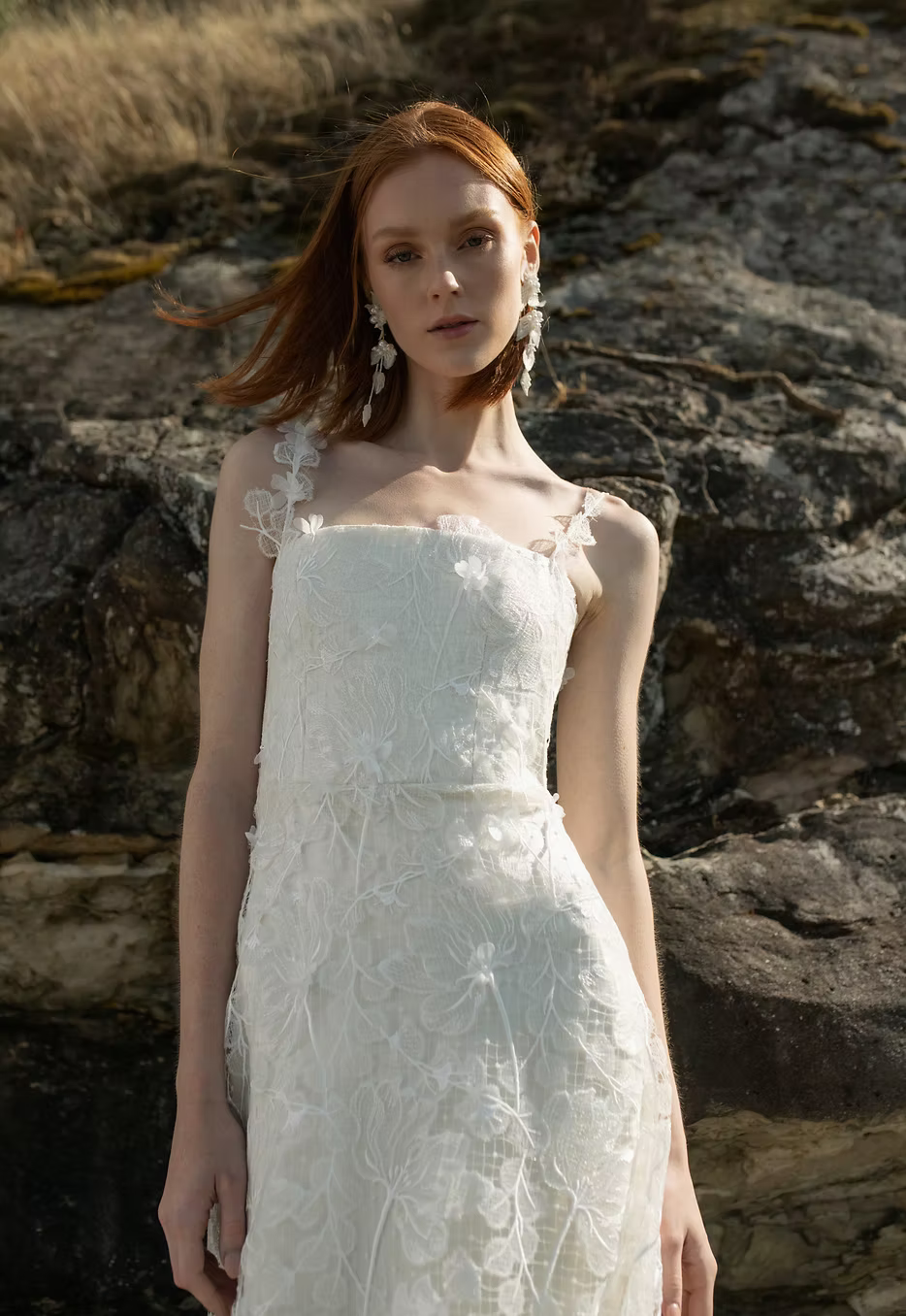 A woman with red hair wearing a white floral lace dress stands outdoors against a rocky, grassy background.