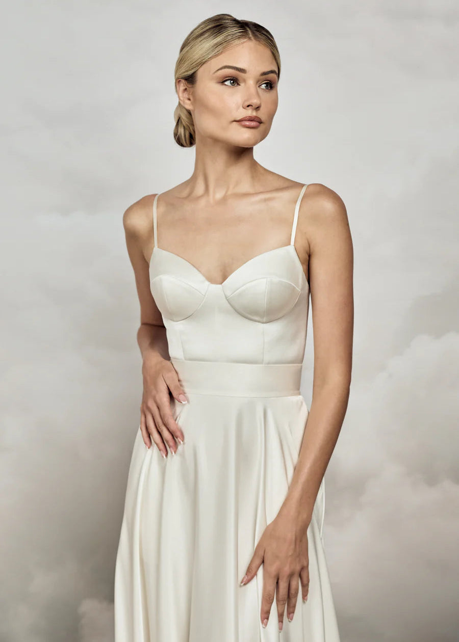 A woman modeling a sleeveless white midi dress with structured underbust cups, thin shoulder straps, and a flowing skirt against a soft gray studio backdrop.