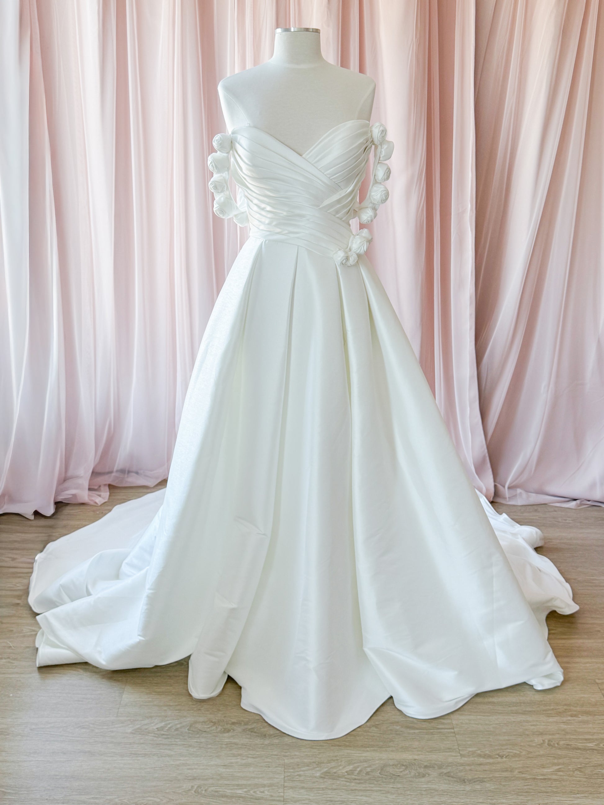 A white strapless wedding gown displayed on a mannequin against soft pink draped curtains and a light wood floor at NKIN. Showing the removable floral rose sleeves and add on waist embellishment.