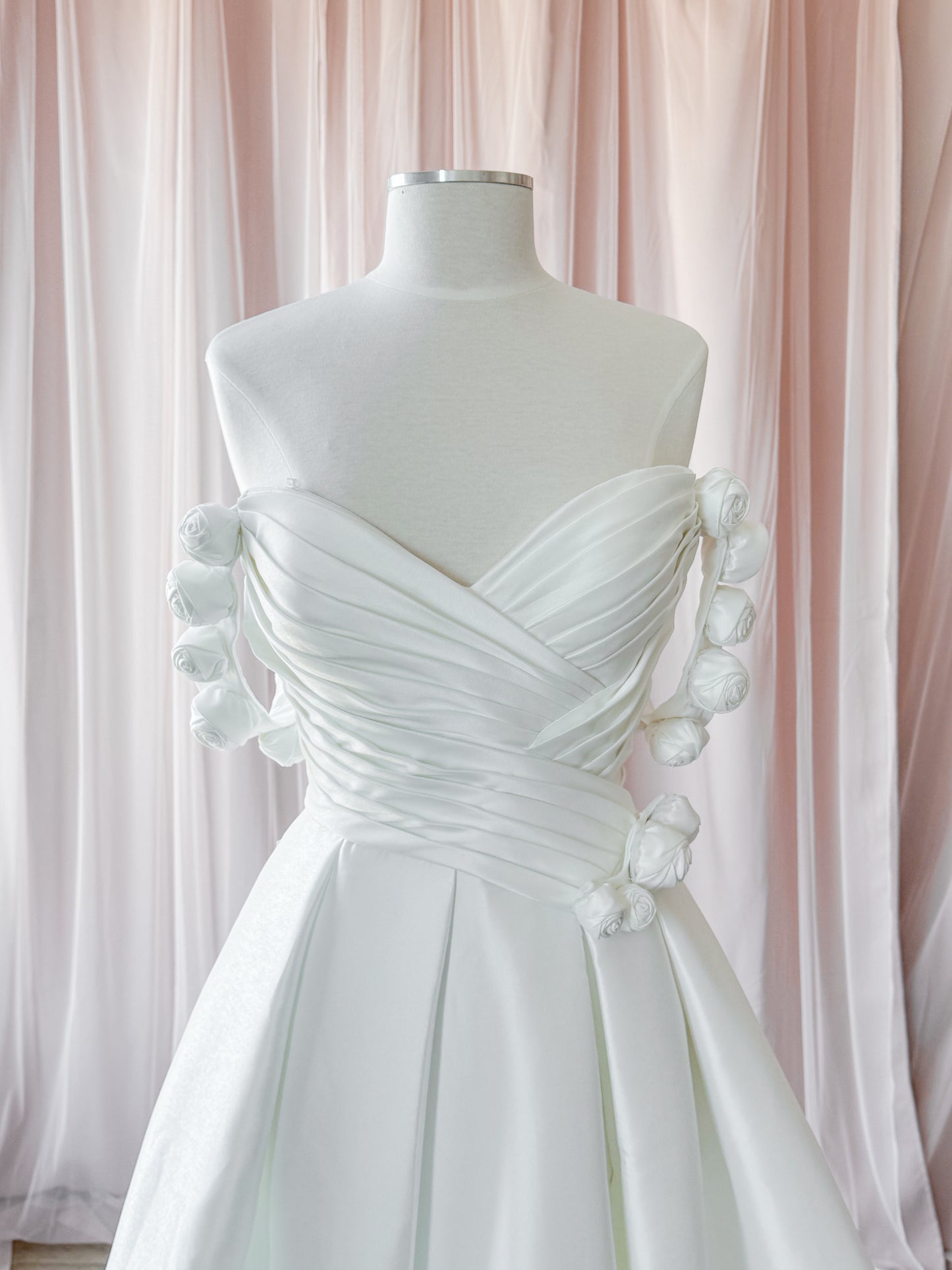 A white strapless wedding dress with ruched draped fabric and basque waist offset. This shows the optional  decorative fabric flowers on the bodice on the waistline and the removable off the shoulder sleeves. Displayed on a white mannequin against a soft pink draped fabric backdrop at NKIN