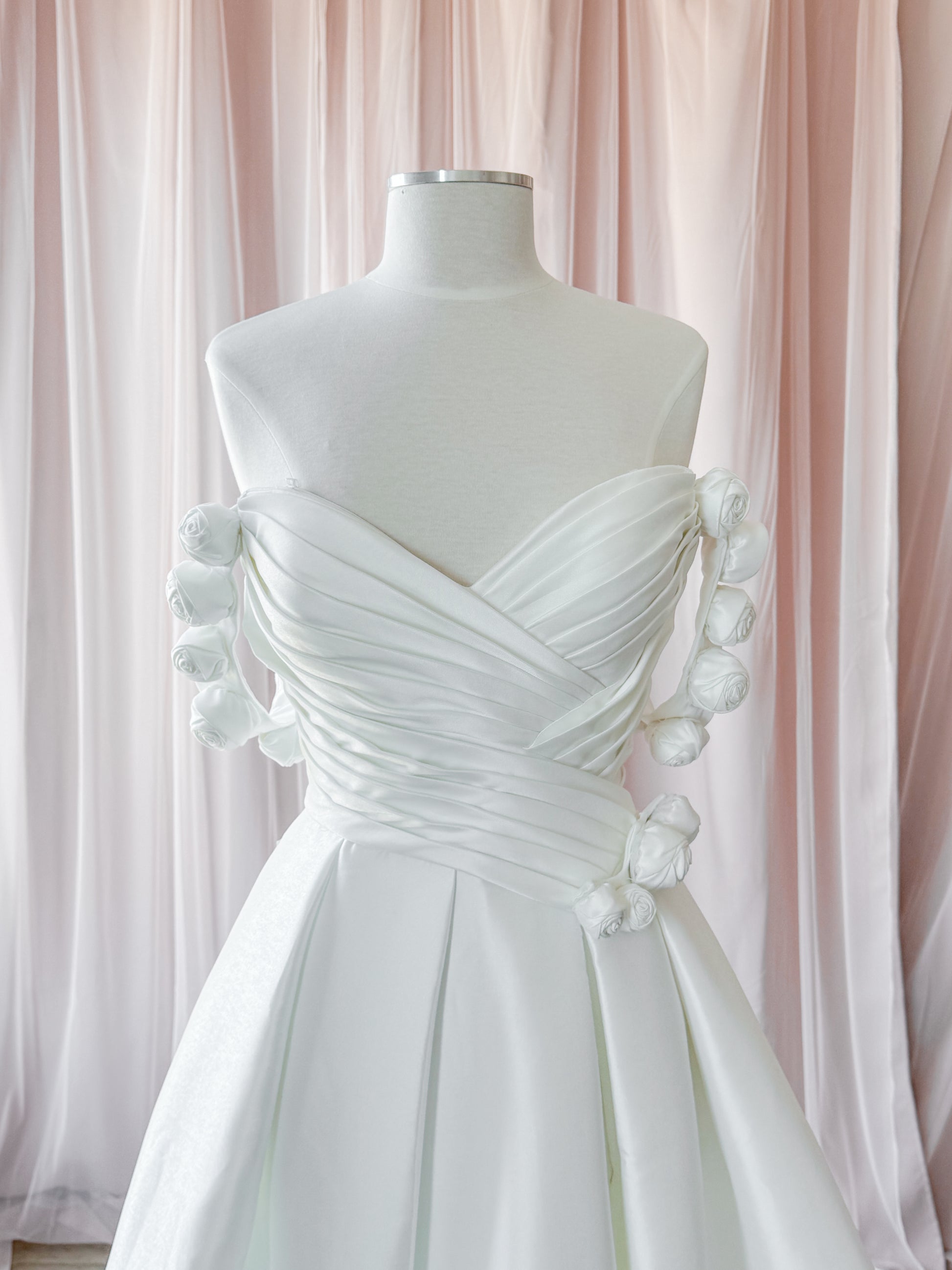 A white strapless wedding dress with ruched draped fabric and basque waist offset. This shows the optional  decorative fabric flowers on the bodice on the waistline and the removable off the shoulder sleeves. Displayed on a white mannequin against a soft pink draped fabric backdrop at NKIN
