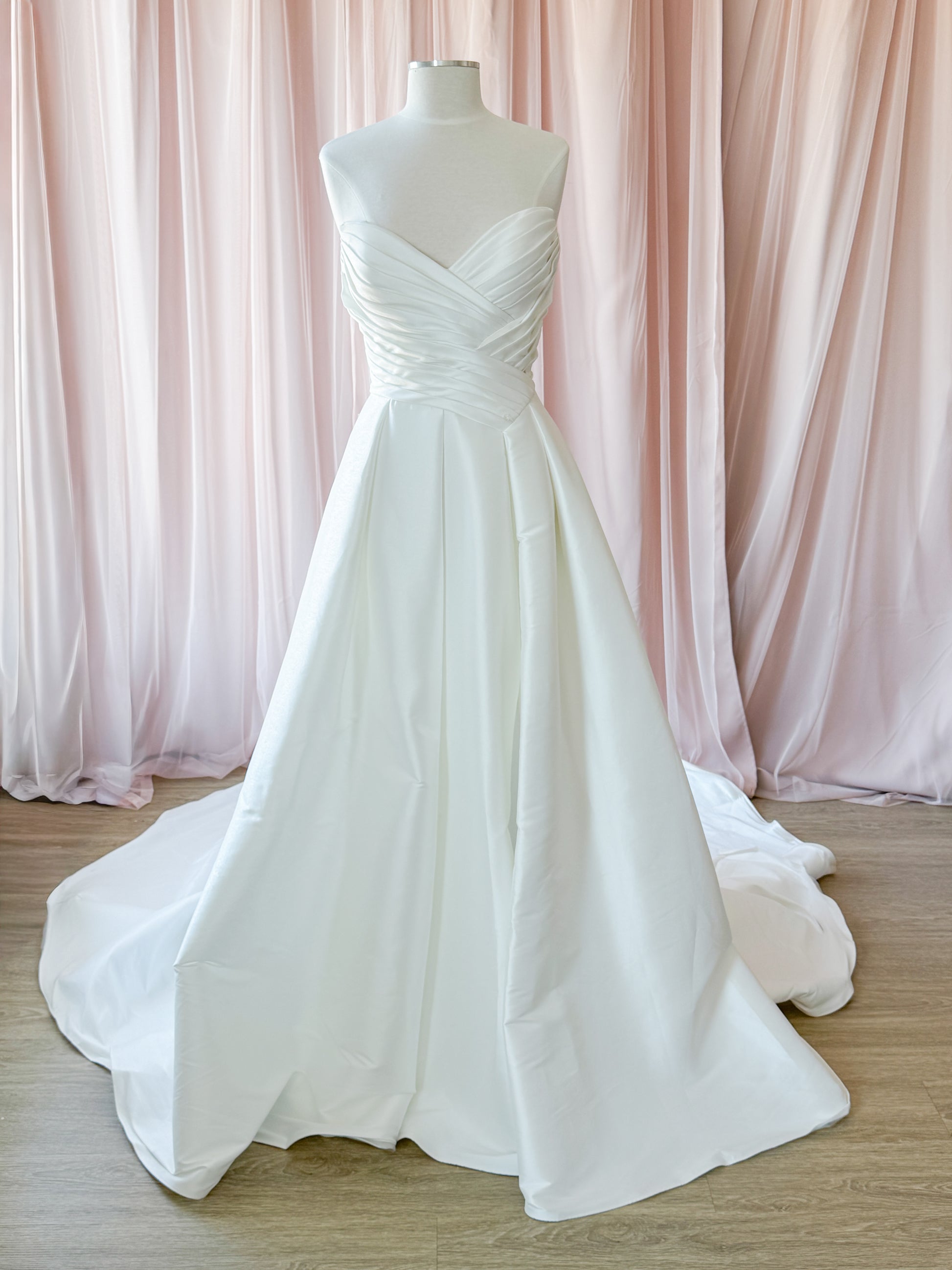 A strapless white wedding gown displayed on a mannequin, featuring a sweetheart neckline, ruched bodice,offset basque waisT  and full A-line skirt with a train, set against pink draped fabric and a light wood floor At Never Knew I Needed Bridal Outlet Ottawa