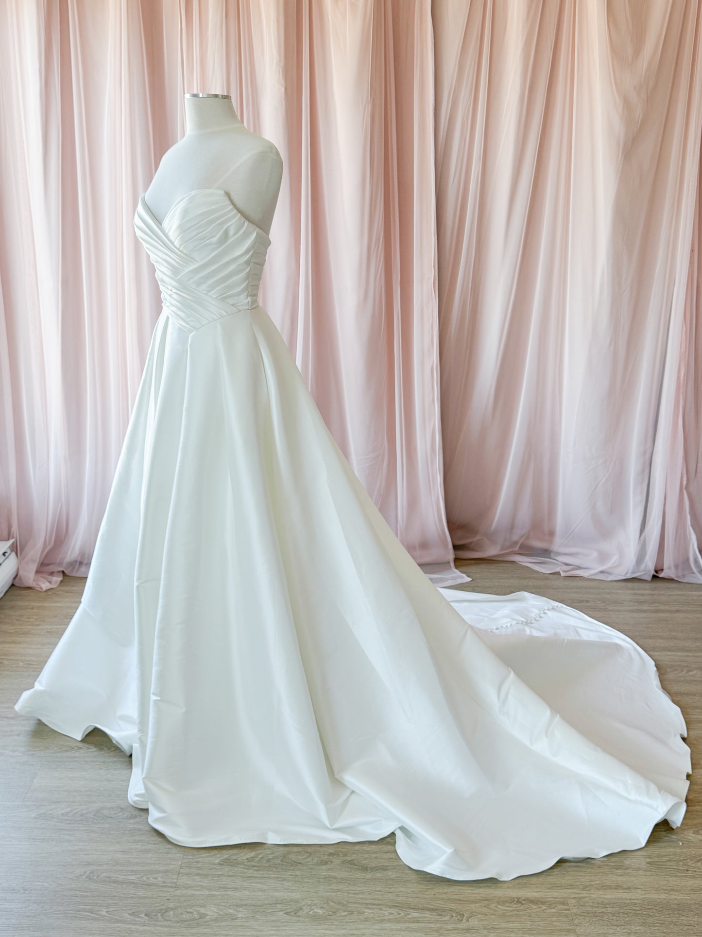 A white mikado A-line wedding gown displayed on a mannequin, featuring a strapless sweetheart neckline, ruched bodice, and a long train, set against a backdrop of draped blush pink fabric in the fitting room at NKIN.
