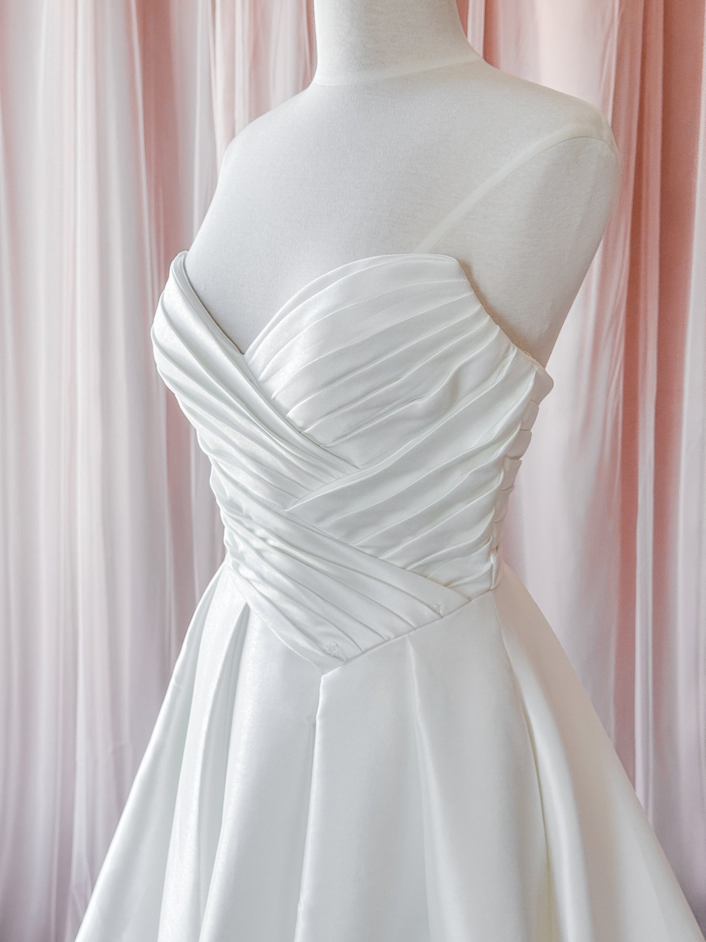 White strapless satin wedding dress on a mannequin, featuring a ruched sweetheart bodice and full skirt, set against a soft pink draped fabric background in a fitting room at clearance bridal shop never knew i needed