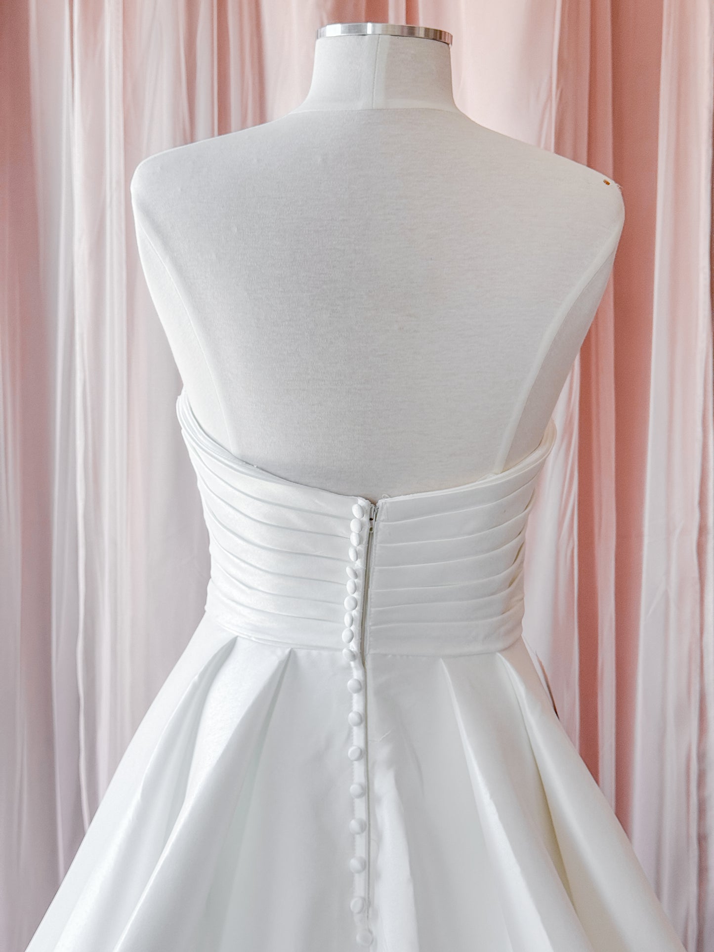 White strapless wedding dress shown from the back on a mannequin, featuring a structured corset-style waist with a central button closure and a flared skirt, set against soft pink draped fabric at NKIN