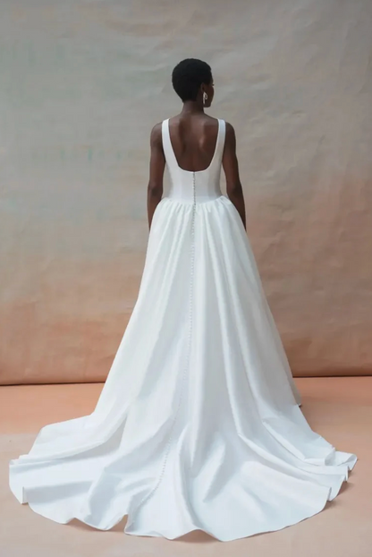 A model viewed from behind wearing a white sleeveless wedding gown with a deep V back and a long train, standing against a warm beige studio backdrop.