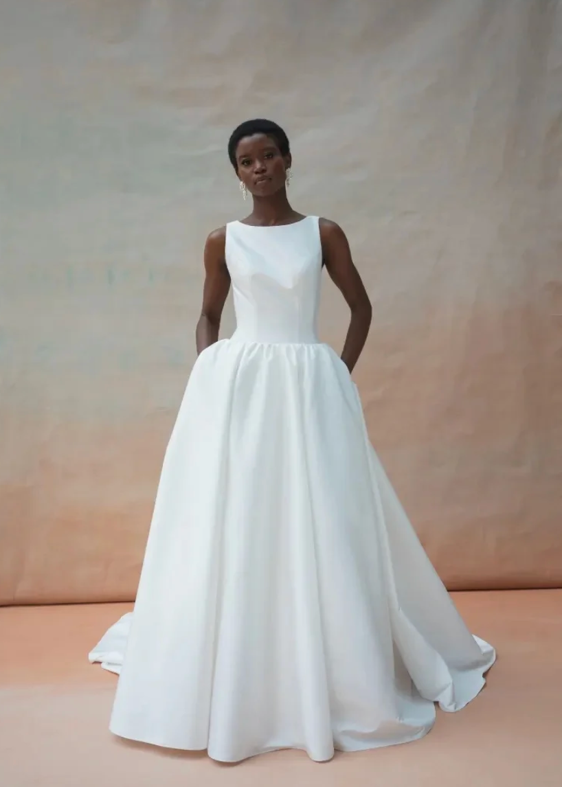 A woman modeling a sleeveless white wedding gown with a structured bodice and a full, flowing skirt against a warm beige studio backdrop.