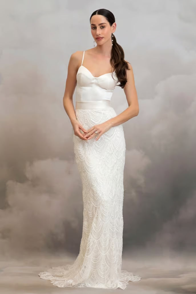A woman modeling a fitted white lace wedding gown with thin straps, a sweetheart neckline, a defined waistband, and a slight train, posed against a smoky gray studio backdrop.