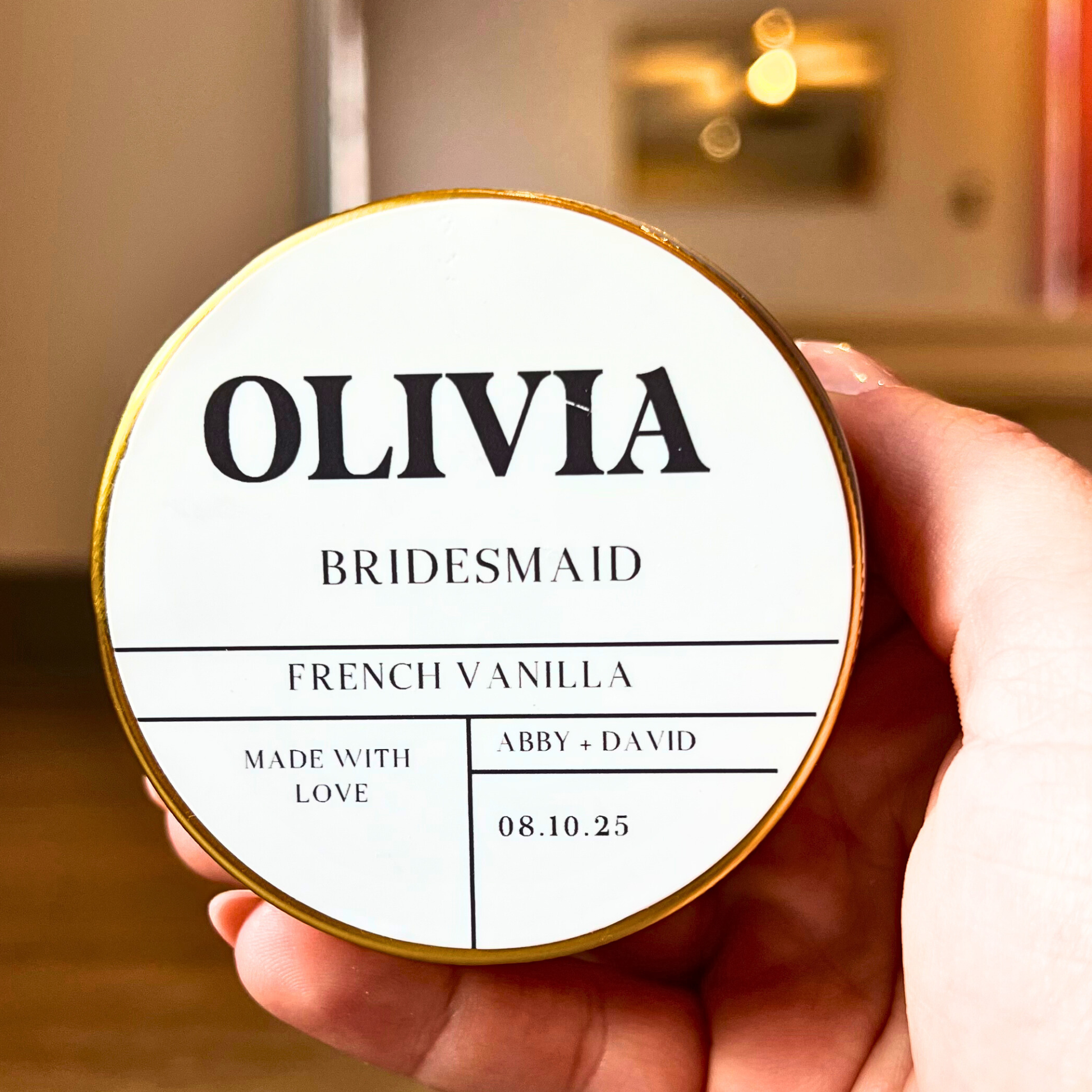 Round container labeled 'OLIVIA BRIDESMAID' held by a hand with a blurred background.