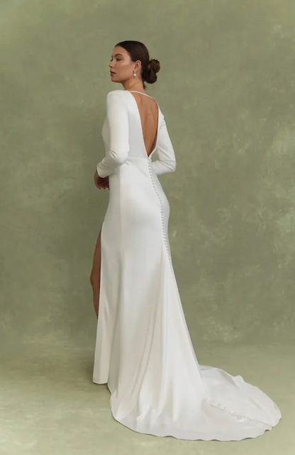 A woman modeling a long-sleeve white wedding gown from the back, showing an open back with decorative buttons and a high slit, against a textured sage green studio backdrop.