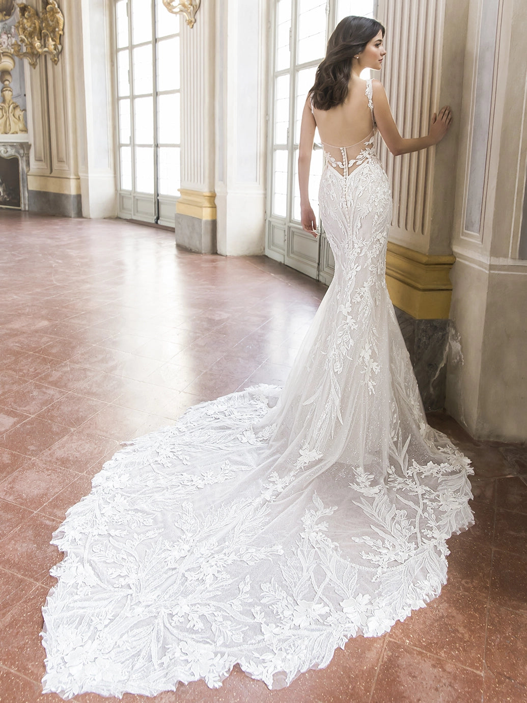 Woman in a white lace wedding dress standing in an elegant room with large windows.