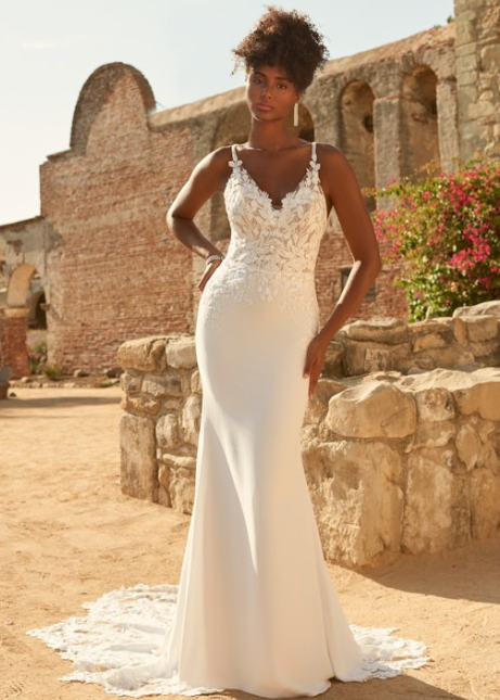 A woman modeling a white sleeveless mermaid-style wedding gown with lace applique and a train, standing outdoors in front of rustic stone and brick ruins.