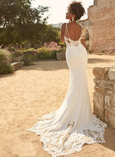 A woman wearing a white lace wedding dress with thin straps and a fitted silhouette, photographed outdoors in a sunlit courtyard with stone walls and trees in the background.