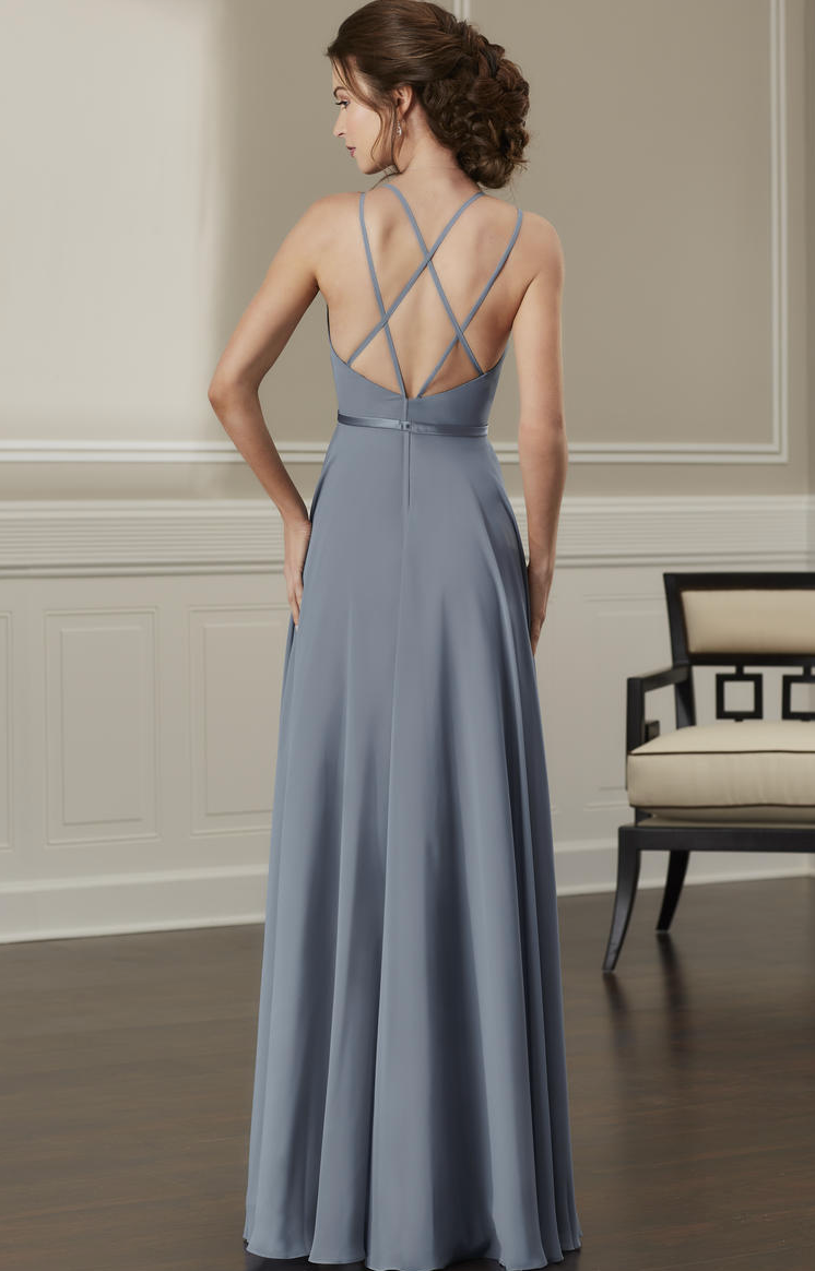 A woman wearing a floor-length dusty blue evening gown, shown from the back in an elegant indoor setting with a paneled wall and a chair.