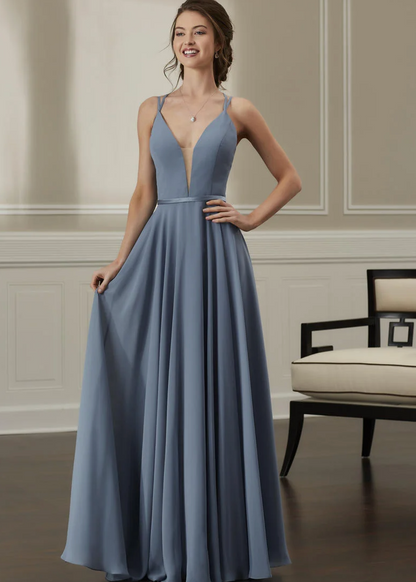A woman modeling a floor-length dusty blue gown with a plunging V-neckline, thin shoulder straps, a defined waist, and a flowing skirt in an indoor room with paneled walls and a chair.