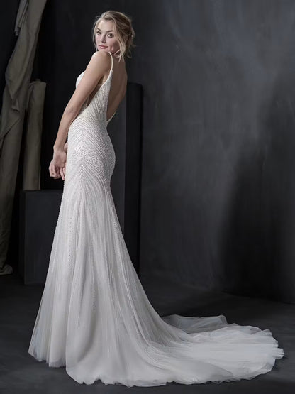A woman modeling a fitted white wedding gown with a long train in a dark studio setting. The dress has thin straps, a fitted silhouette, and a sheer tulle train.