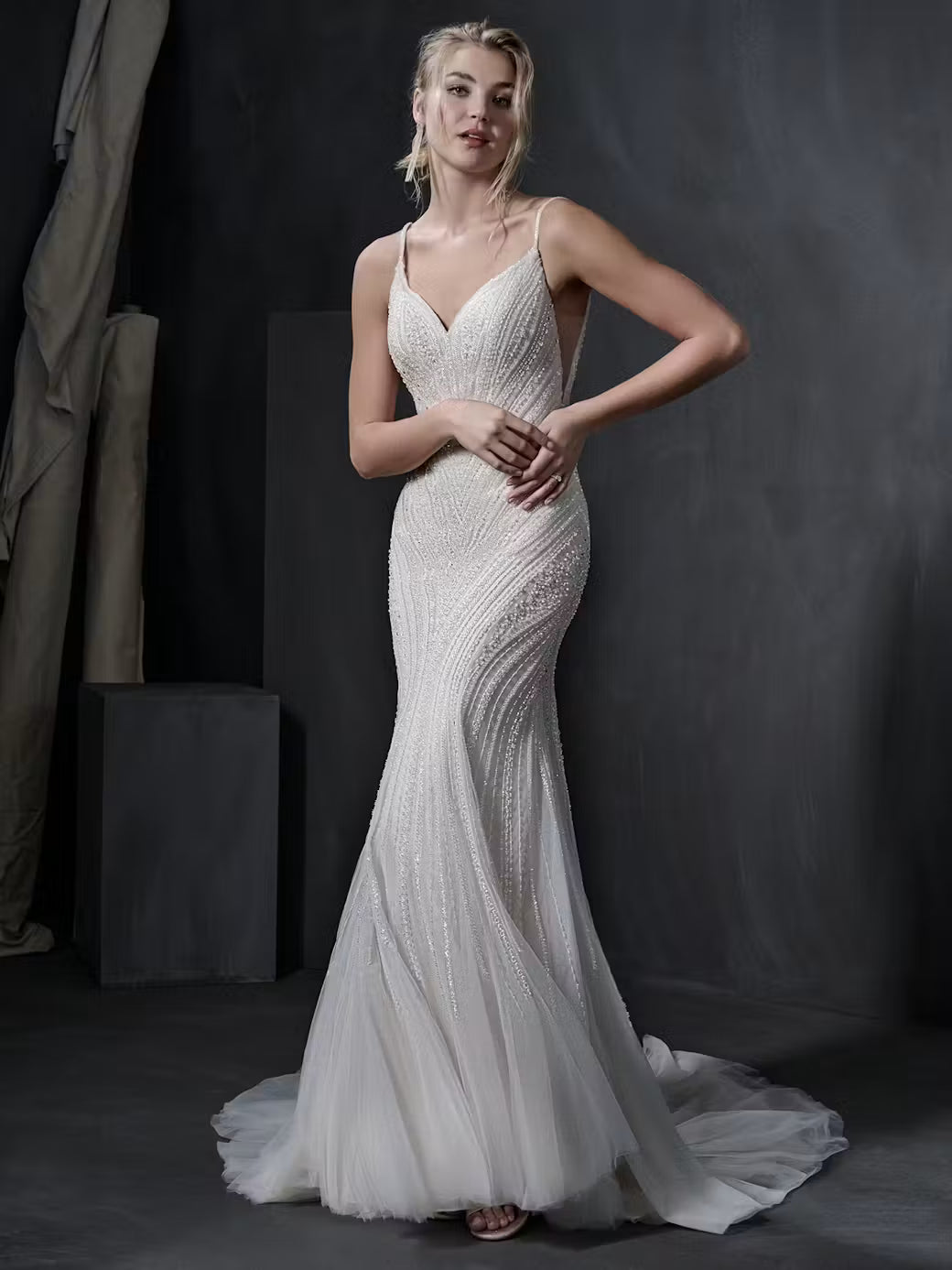 A woman modeling a fitted champagne wedding gown with beaded texture, thin straps, a deep V-neckline, and a flowing tulle train against a dark studio backdrop.