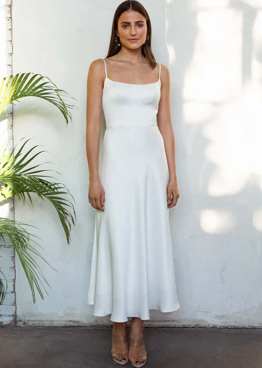 Woman wearing a white dress standing against a white wall with a plant to the left.