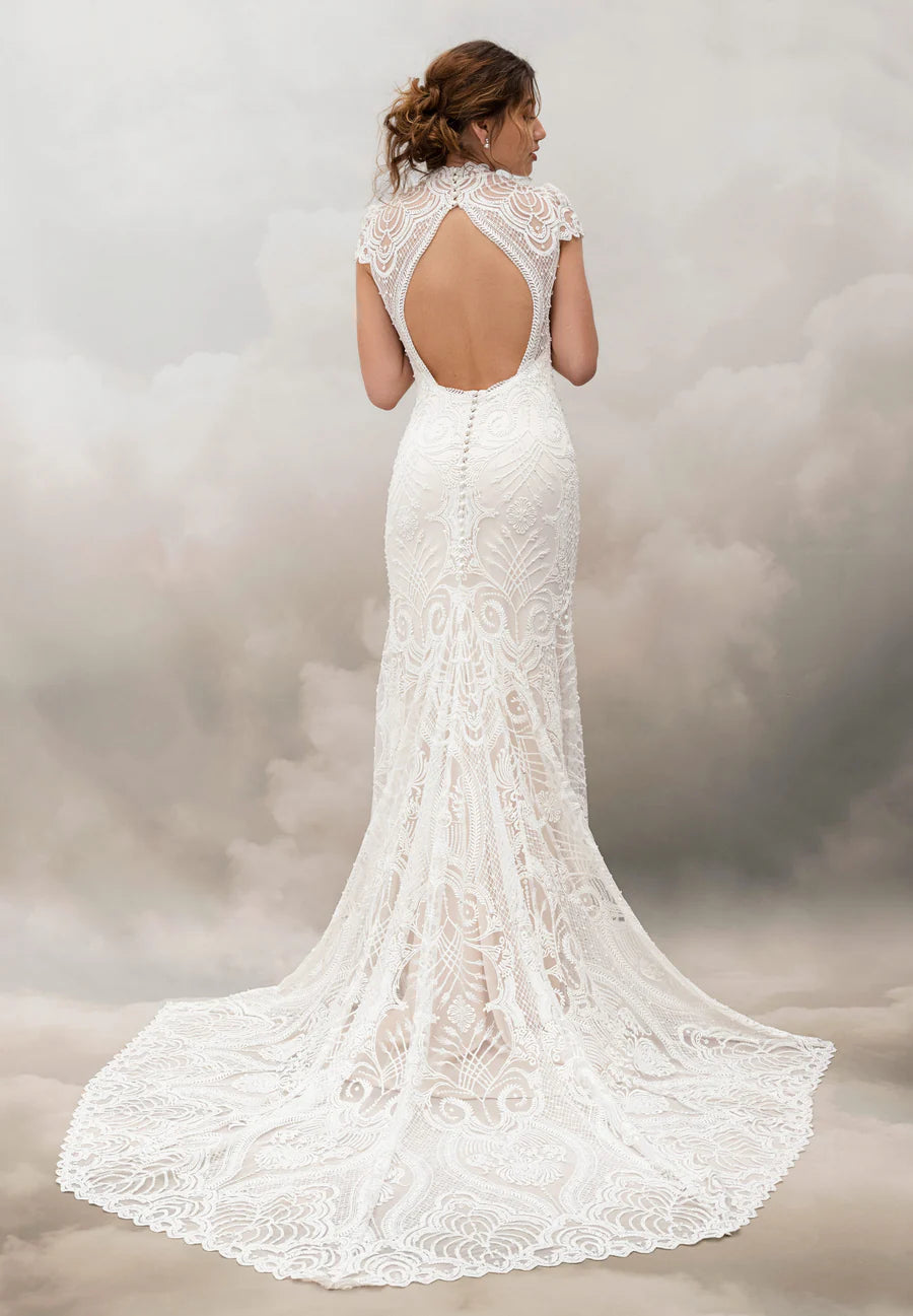 A woman shown from the back wearing a white lace bridal gown with a fitted silhouette, short lace sleeves, an open back, and a long train against a soft gray studio backdrop.