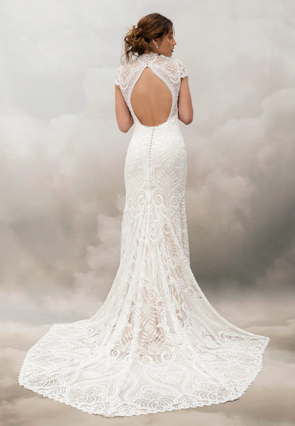A woman shown from the back wearing a white lace bridal gown with a fitted silhouette, short lace sleeves, an open back, and a long train against a soft gray studio backdrop.