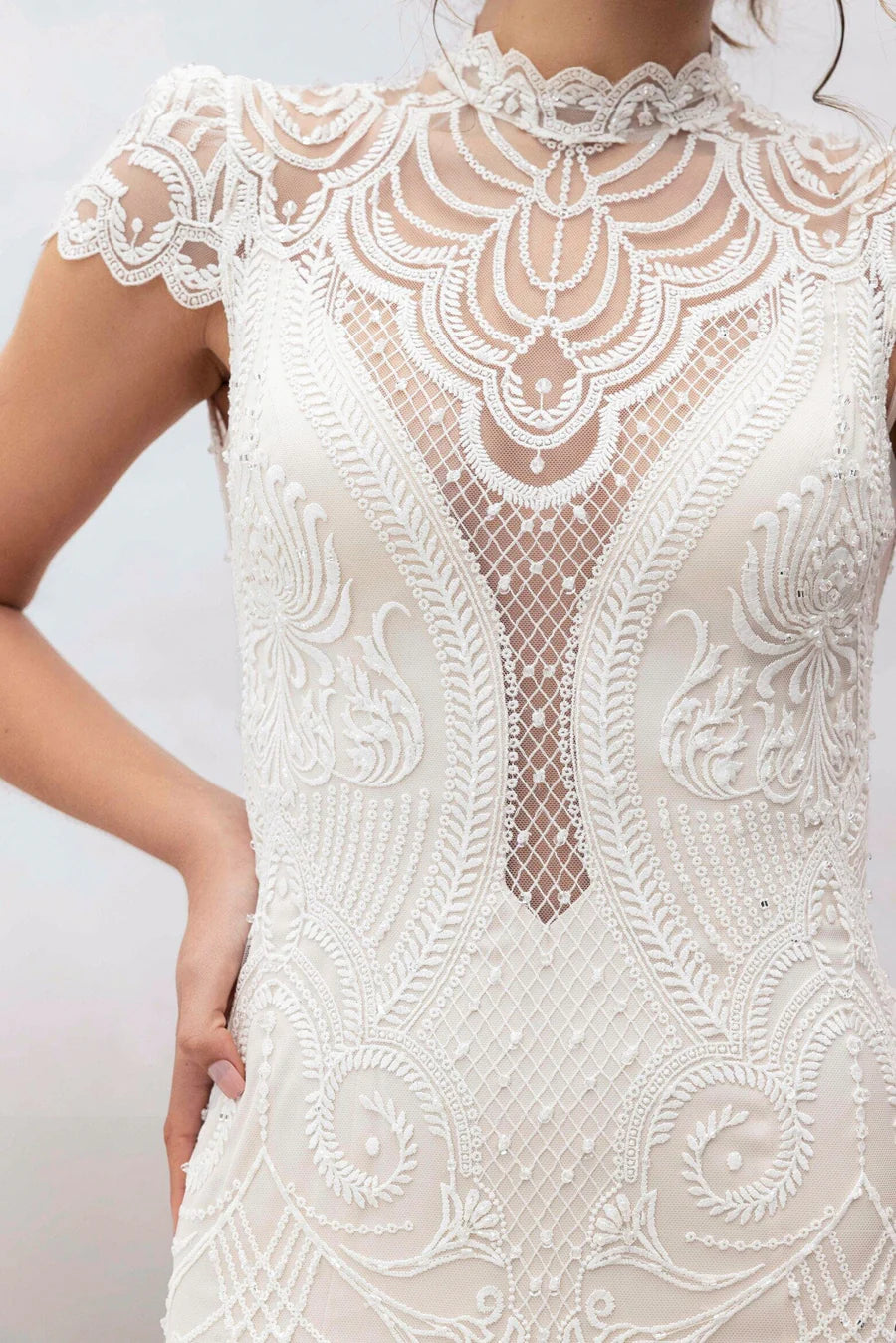 A close-up of a white lace and mesh dress with intricate floral embroidery, a high scalloped neckline, and short cap sleeves, shown on a model against a light gray background.