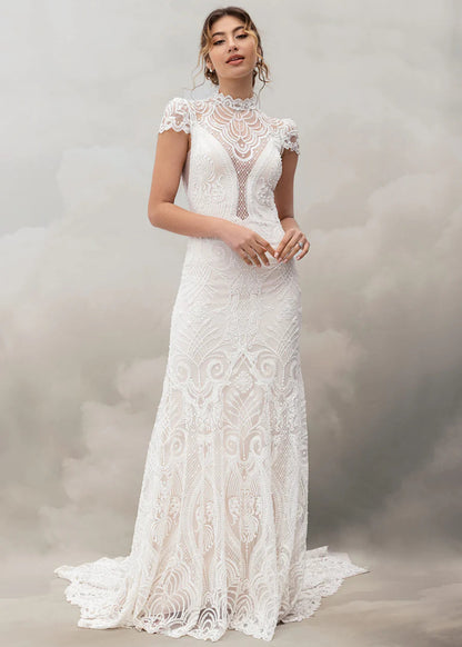 A woman modeling a white lace wedding gown with a high mock neck, cap sleeves, and a fitted mermaid silhouette with a slight train, posed against a soft gray studio backdrop.
