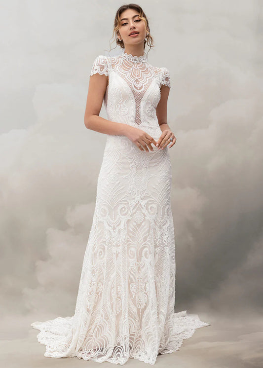 A woman modeling a white lace wedding gown with a high mock neck, cap sleeves, and a fitted mermaid silhouette with a slight train, posed against a soft gray studio backdrop.