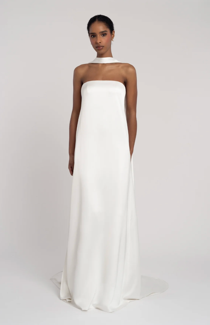 A woman modeling a strapless white satin gown with a high halter-style neckline and a long flowing skirt, photographed against a plain light gray studio background.