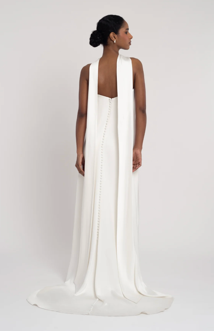 A woman modeling a floor-length white gown from the back, shown against a plain light gray studio background. The dress has a deep open back, a long draped shawl-like back panel, and a slight train.