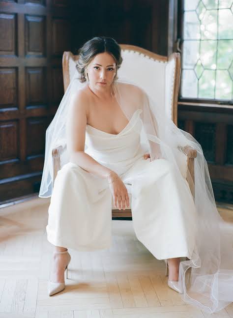 A woman seated in a vintage upholstered armchair wearing a strapless white bridal-style jumpsuit with wide legs and a sheer veil, in an indoor room with dark wood paneling and a window.