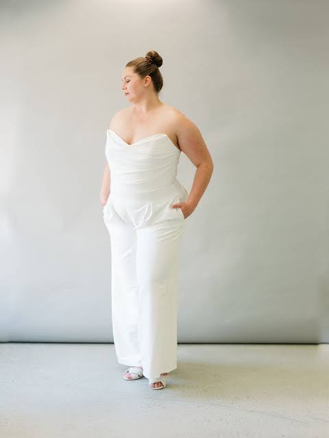 A woman modeling a white strapless jumpsuit with wide-leg pants, standing against a neutral light gray studio backdrop.