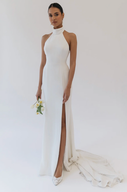 A woman stands in a studio wearing a floor-length white halter gown with a high thigh slit and a long train. She is holding a small bouquet of white flowers and wearing white pointed shoes against a plain light background.