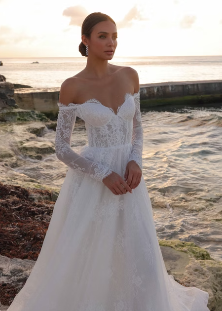 Woman in a white lace wedding dress standing by the ocean at sunset.