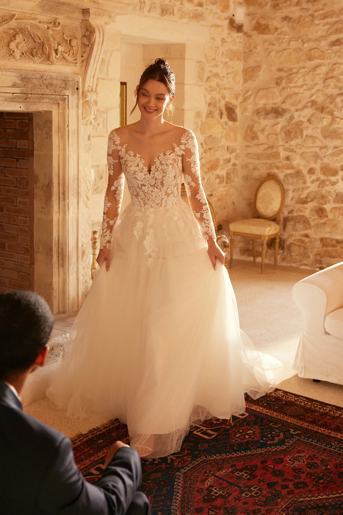 A woman wearing a long-sleeve lace wedding gown with a full tulle skirt stands in a warmly lit stone room, with a seated man partially visible in the foreground.
