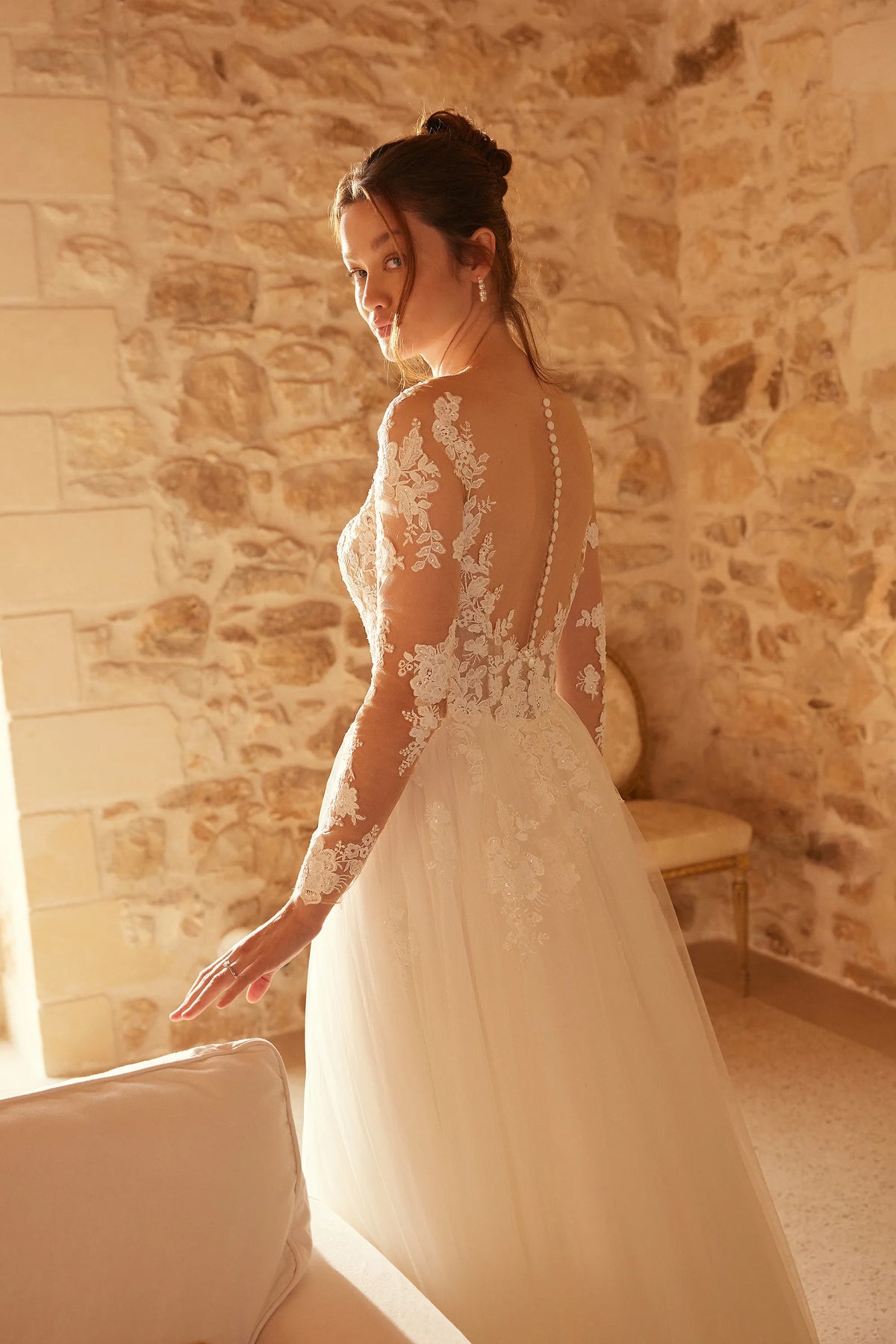 A woman wearing a long-sleeved ivory lace wedding gown with a sheer back, standing in a warm-lit room with a stone wall background.