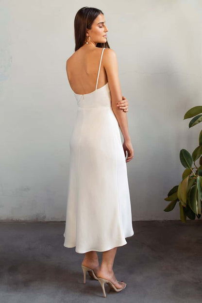 Woman in a white dress standing against a plain wall with a plant to the side.