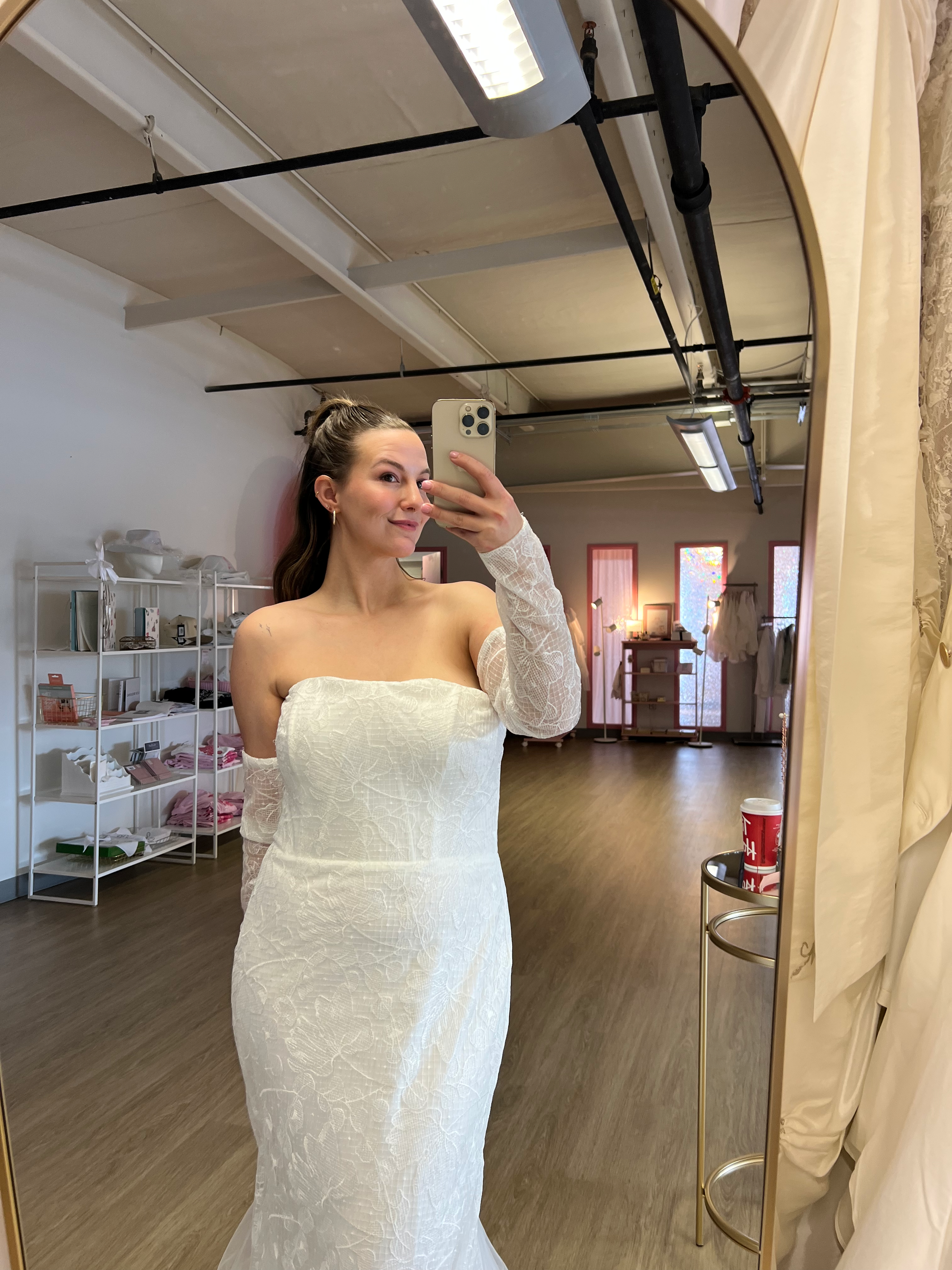 Abby taking a mirror selfie at NKIN, off the rack bridal shop, wearing a white strapless dress , the Elvira, in the NKIN showroom 