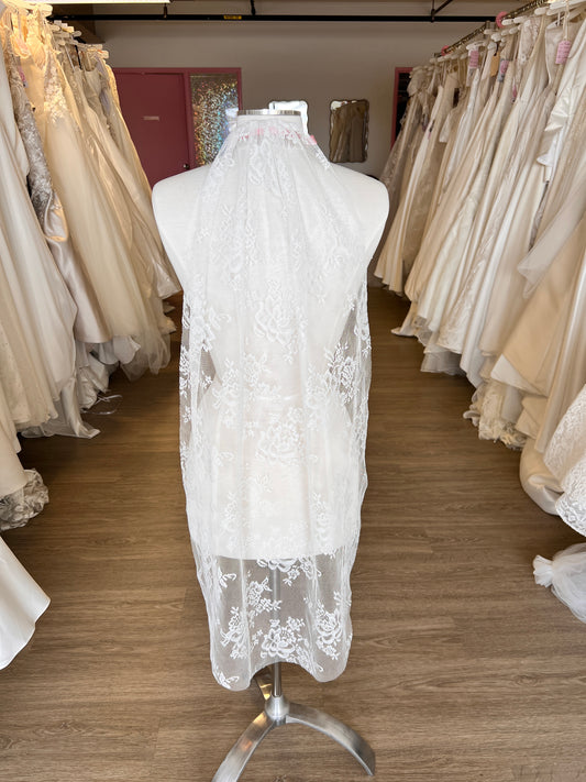 White lace wedding dress on a mannequin in a store setting with other dresses stacked in the background.
