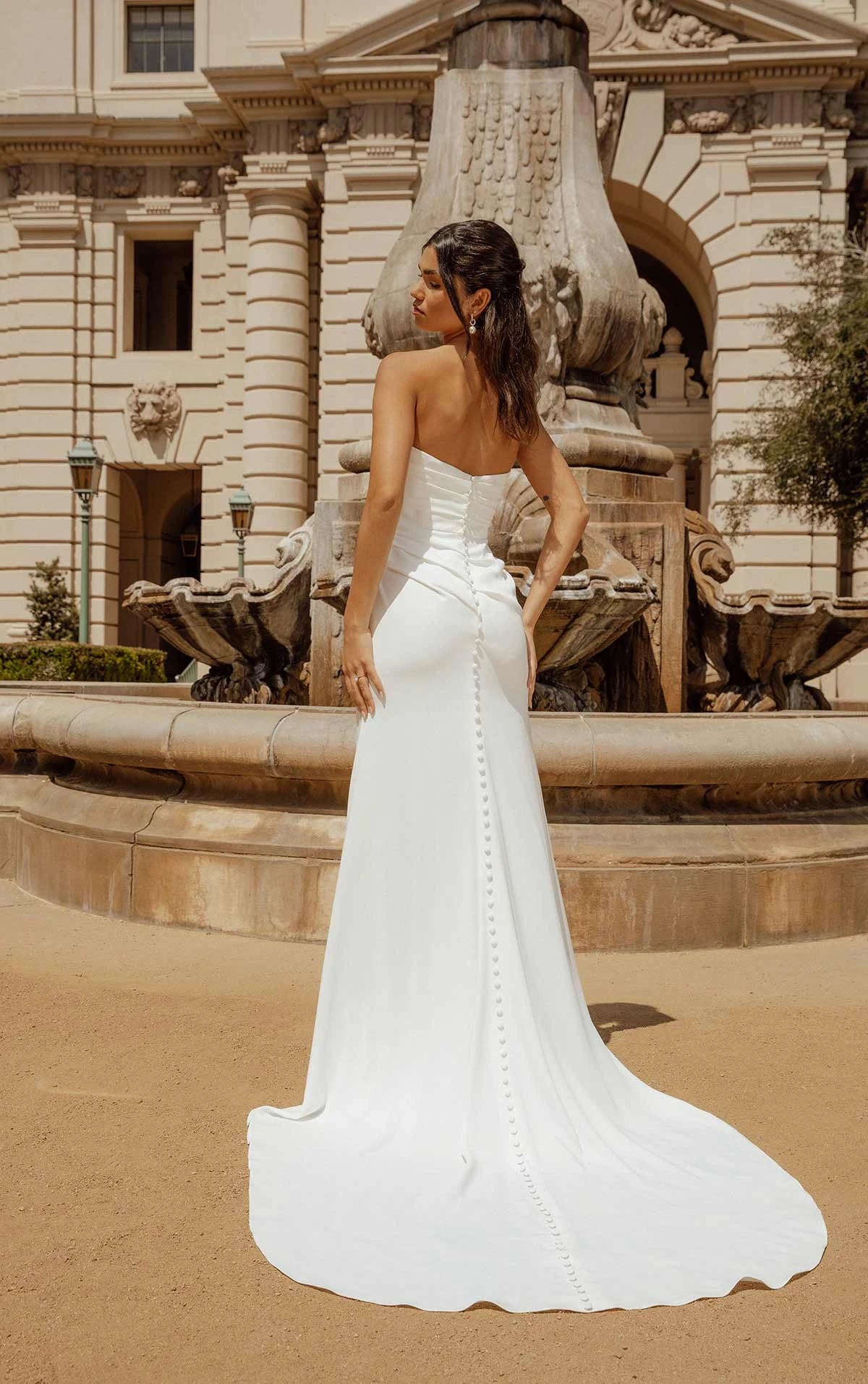 A woman wearing a strapless white wedding gown with a fitted bodice and a long train, photographed outdoors in front of a large stone fountain and classical building.