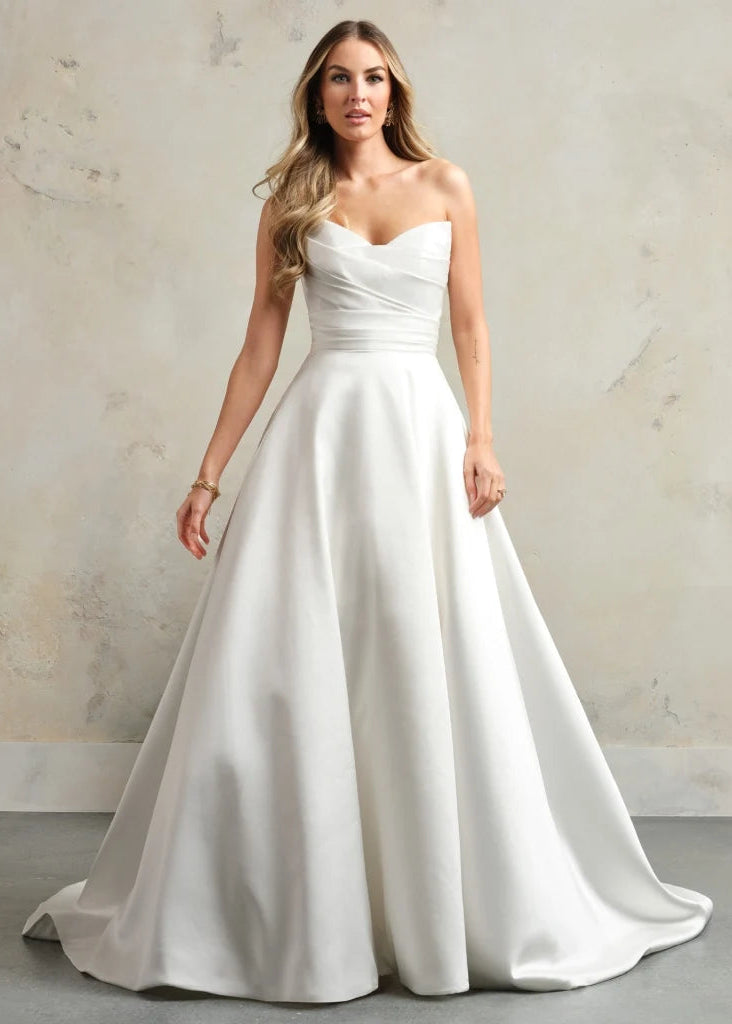 A woman modeling a strapless white A-line wedding gown with a sweetheart neckline, ruched bodice, and full satin skirt against a textured beige studio backdrop.
