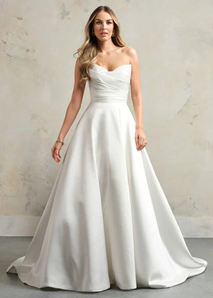 A woman modeling a strapless white A-line wedding gown with a sweetheart neckline, ruched bodice, and full satin skirt against a textured beige studio backdrop.