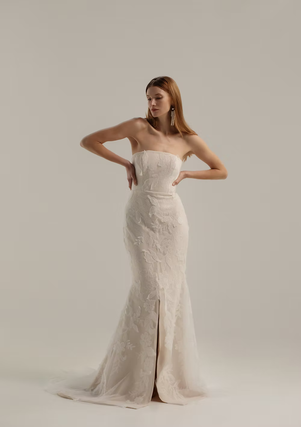 A woman modeling a strapless ivory lace mermaid gown with a thigh-high front slit, posed against a seamless light beige studio background.