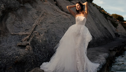 A woman wearing a strapless white tulle wedding gown stands on a rocky shoreline beside water, posing with her hands raised against a large gray rock face.
