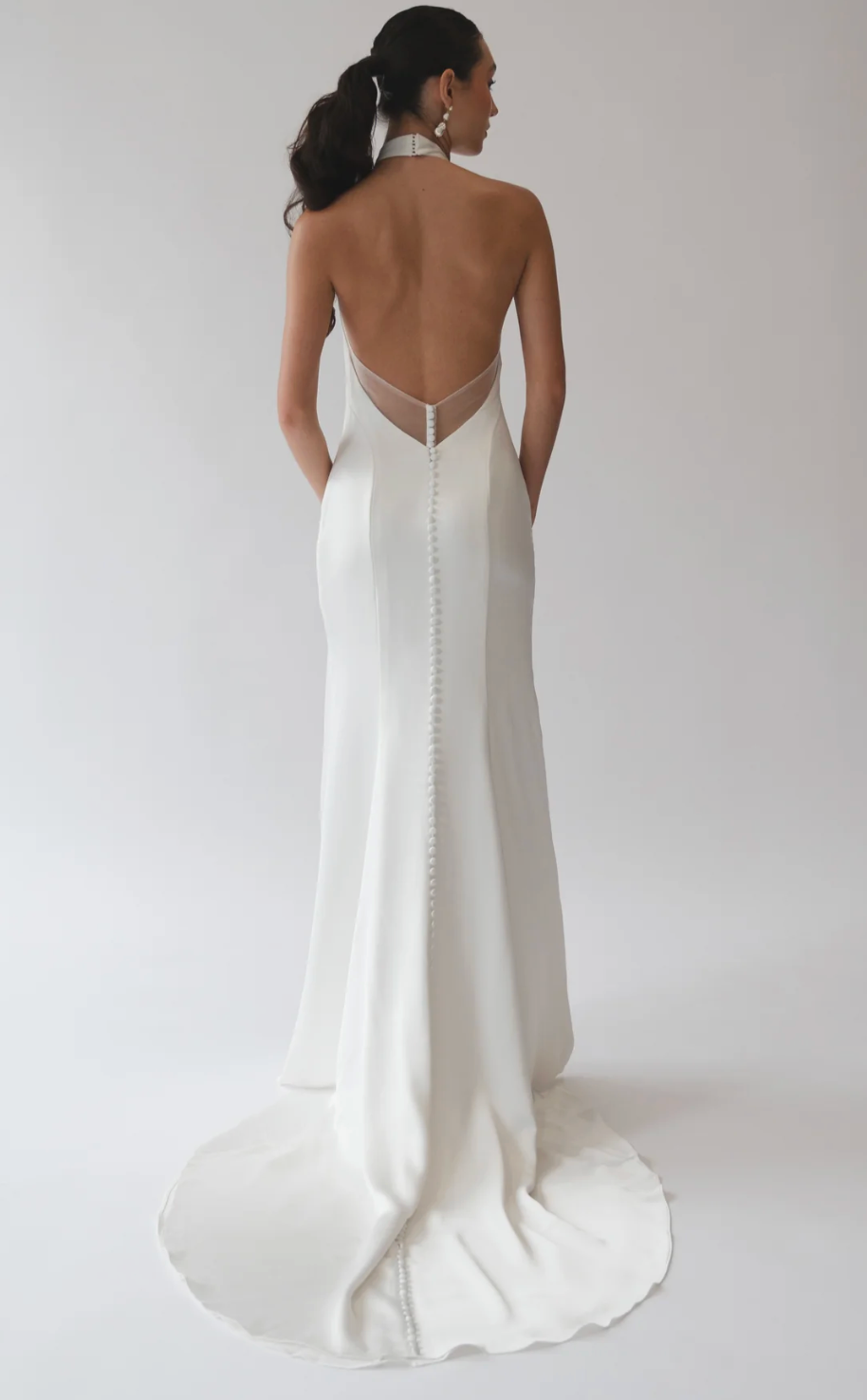 A woman shown from the back wearing a fitted white bridal gown with an open back, sheer mesh panel at the neckline, and a long train on a light gray studio background.